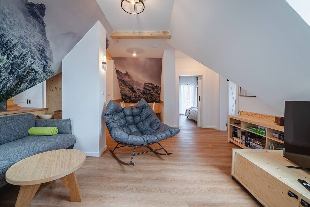 A modern living room with a sofa, a rocking chair, a wooden coffee table, a TV stand, and a bookshelf.