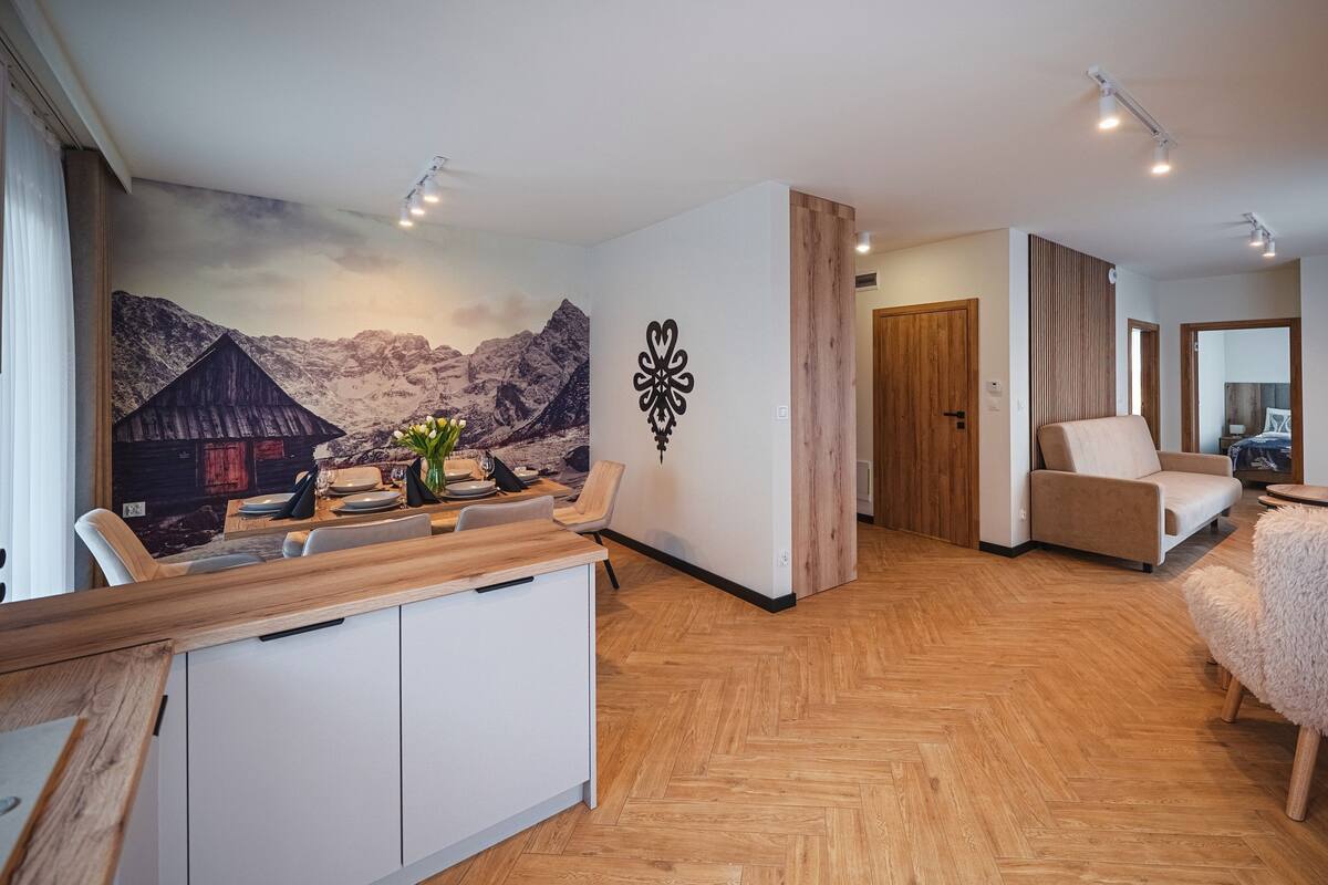 A modern living room with a dining area, wooden herringboard flooring, a large wall mural, and a fluffy chair.