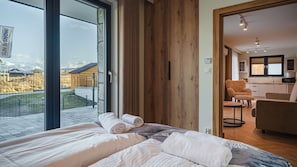 A bedroom with a large window, a wooden wall, and a view of a building and mountains. - SKI Apartments II, Sun&Snow (Białka Tatrzańska)