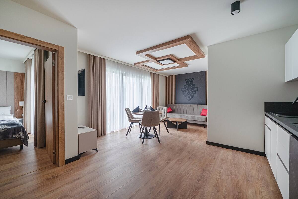 A modern hotel room with a wooden floor, a bed with a headboard, a dining area with a table and chairs, a sofa, and a kitchen area with a sink.
