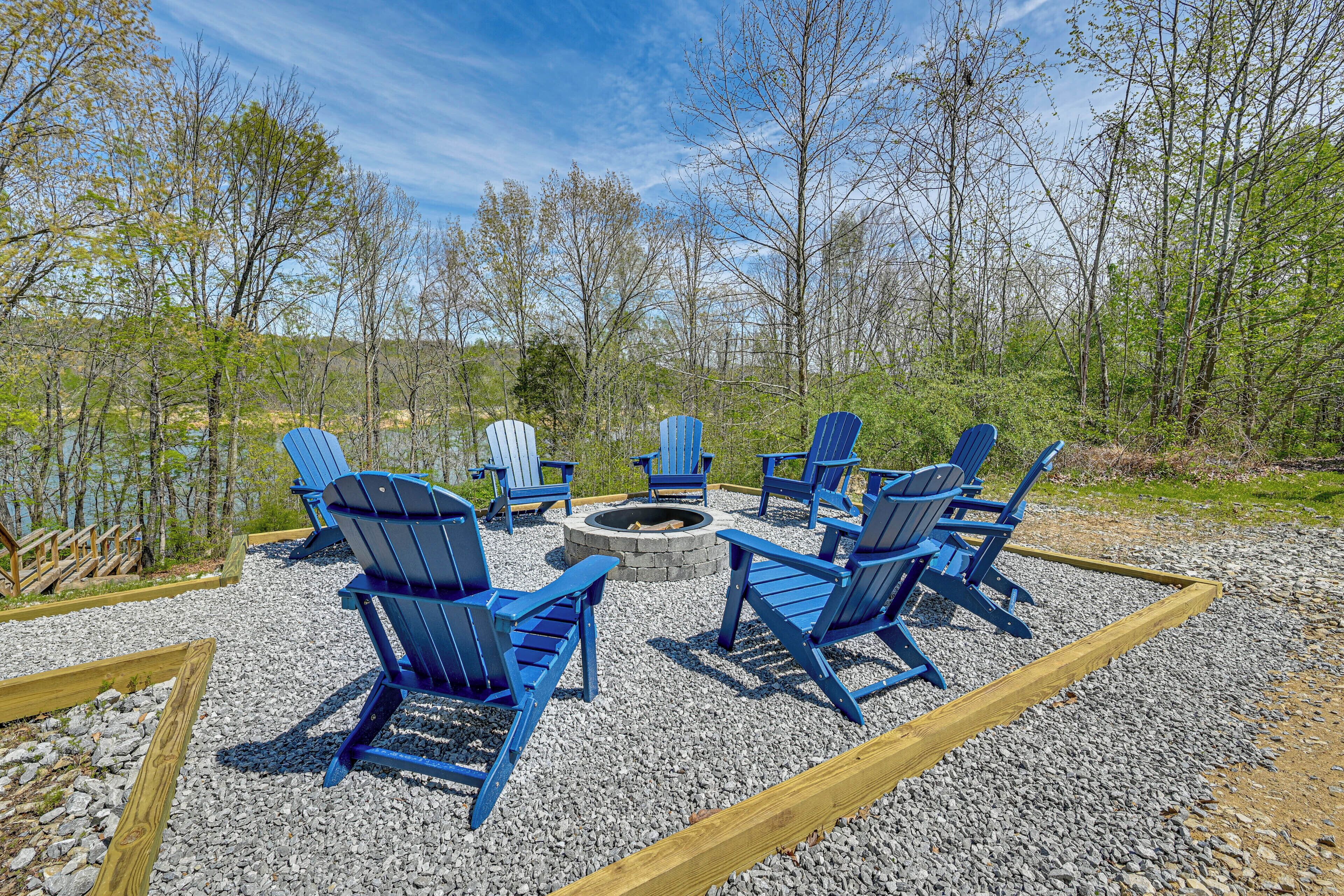 Een terras met Adirondack-stoelen rond een vuurkuil, omgeven door grind en houten randen, met bomen op de achtergrond.