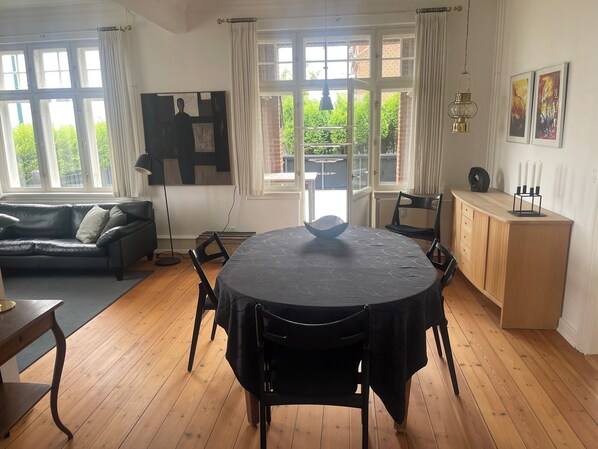 Una habitación con una mesa redonda, sillas negras, un armario de madera, un sofá negro y una ventana con cortinas.