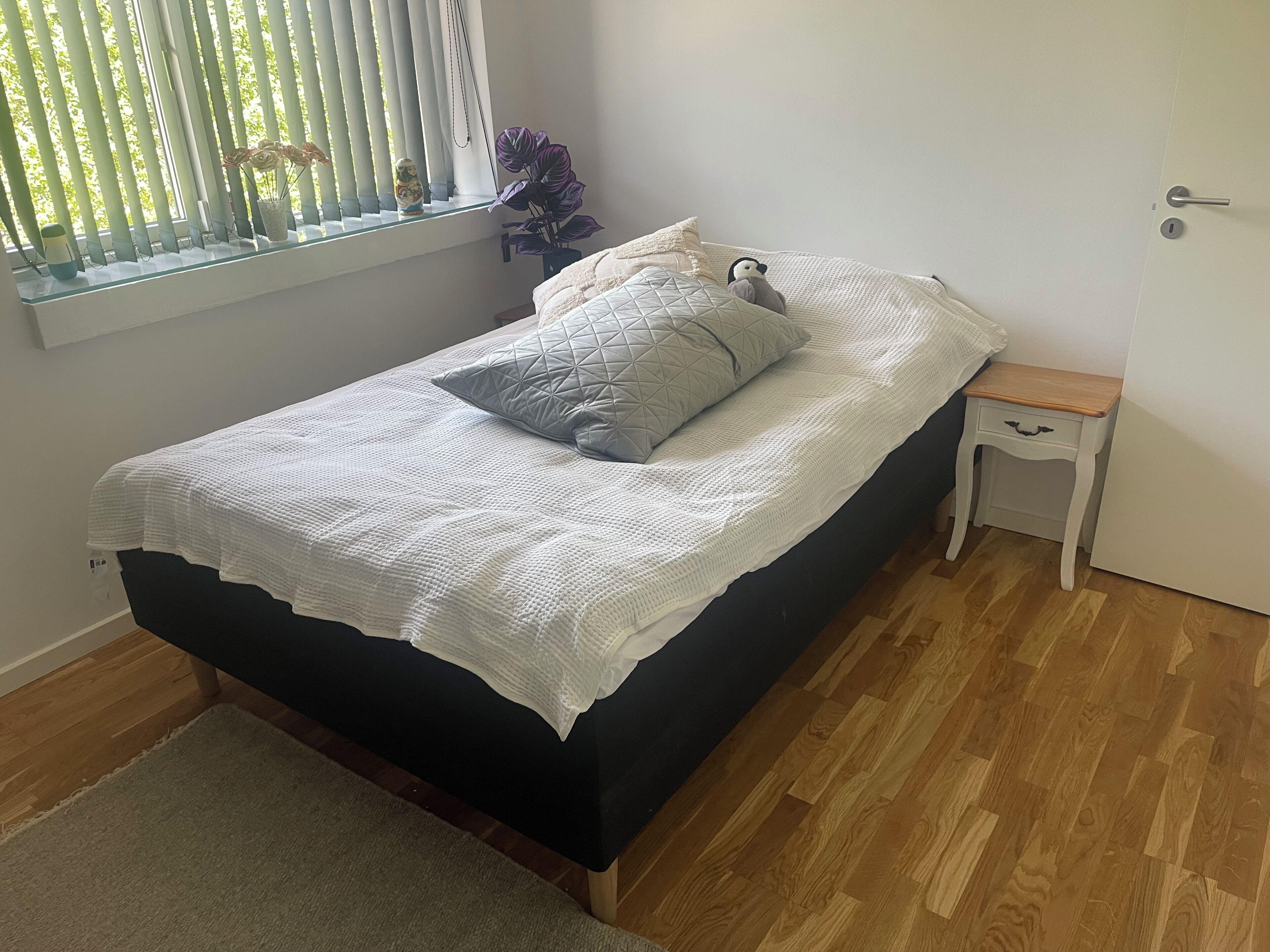 Un lit avec une couette blanche, un oreiller gris et une petite table de chevet dotée d’un tiroir.