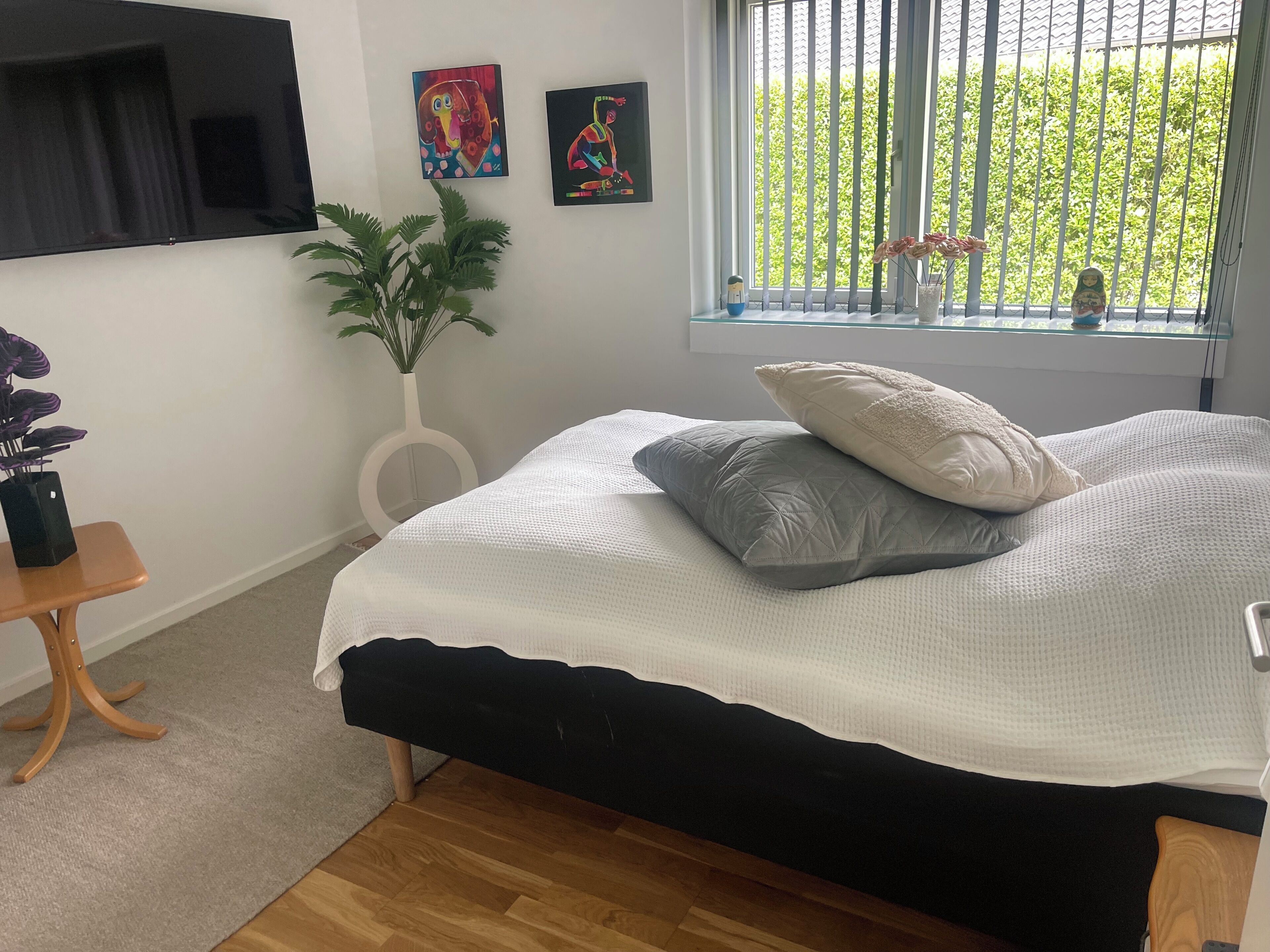 A bedroom with a bed, a potted plant, artwork on the wall, and a window with a view.
