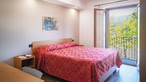 Quarto com cama, escrivaninha, mesa de cabeceira, janela com vista para árvores e um quadro na parede.