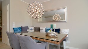 A dining area with a glass-top table, grey upholstered chairs, a decorative chandelier, and a mirror above the table. - Modern 93sqm on 2 levels for up to 4 people with its own sauna - (Sylt)