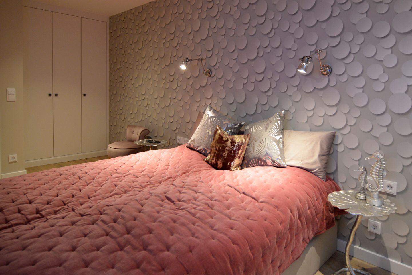 A bedroom with a pink quilted bedspread, a seahorse lamp, and a wall with a circular pattern.