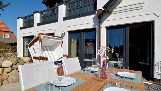 A dining area with a table set for four, outdoor seating, and a white building with balconies. - Modern 93sqm on 2 levels for up to 4 people with its own sauna - (Sylt)
