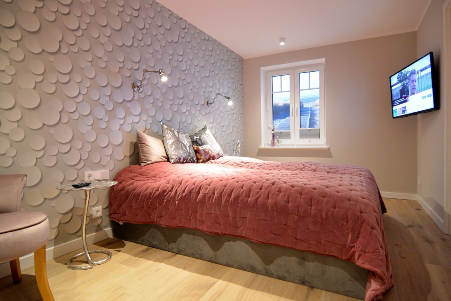 A bedroom with a patterned wall, a bed with a pink blanket, a small table with a lamp, and a TV on the wall.