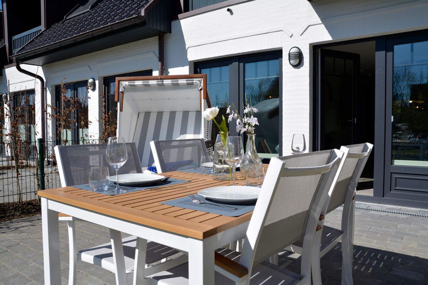 Un'area pranzo all'aperto con un tavolo in legno e sedie bianche, affacciata su un edificio moderno con ampie finestre.