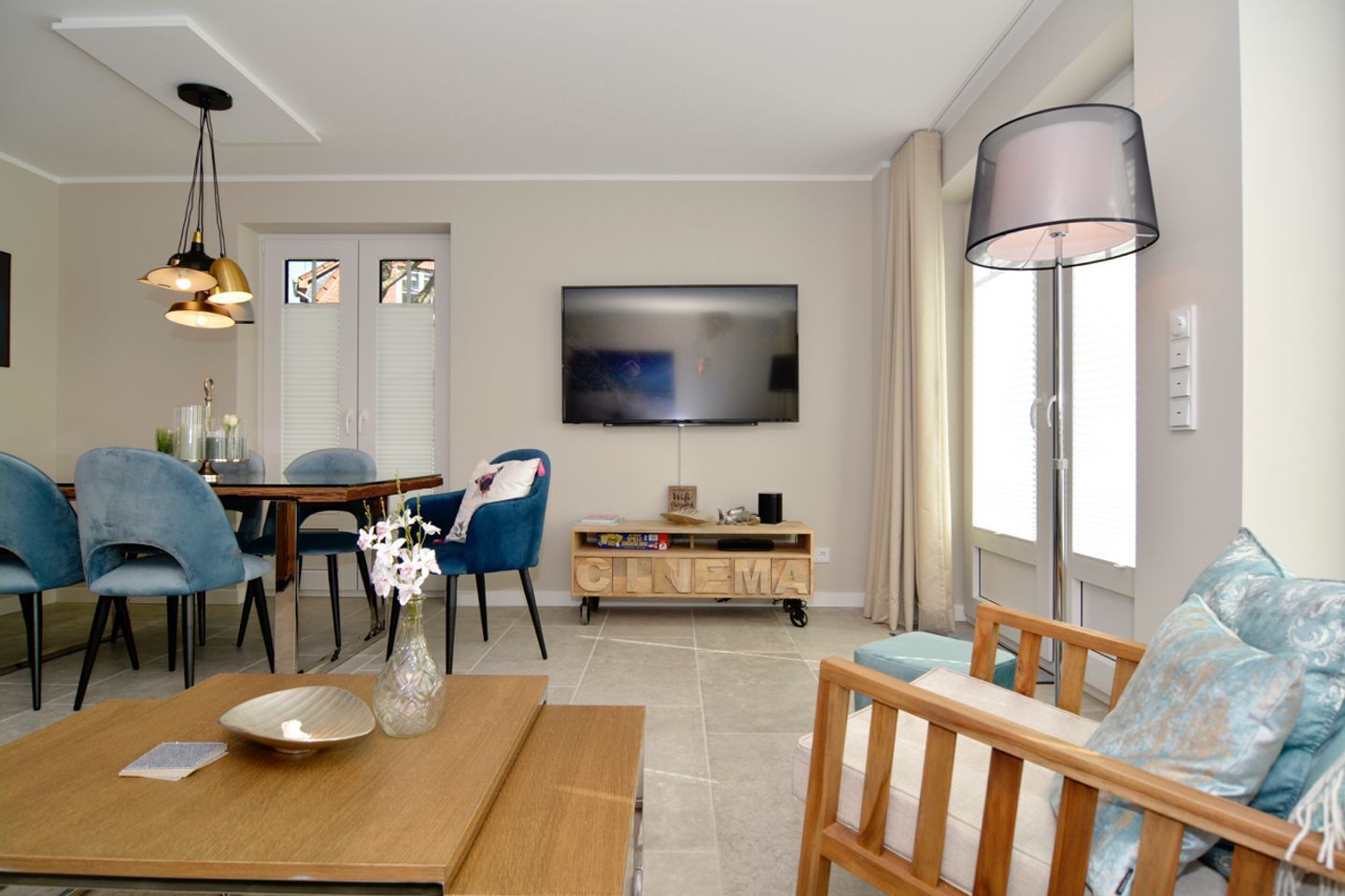 Ein Wohnzimmer mit Fernseher, blauen Sesseln, einem Holztisch, einem TV-Schrank und einem großen Fenster.