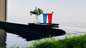 Un bicchiere con liquido rosso e blu su un vassoio nero, un vaso di vetro trasparente con fiori bianchi, e una vista su uno specchio d'acqua e vegetazione.
