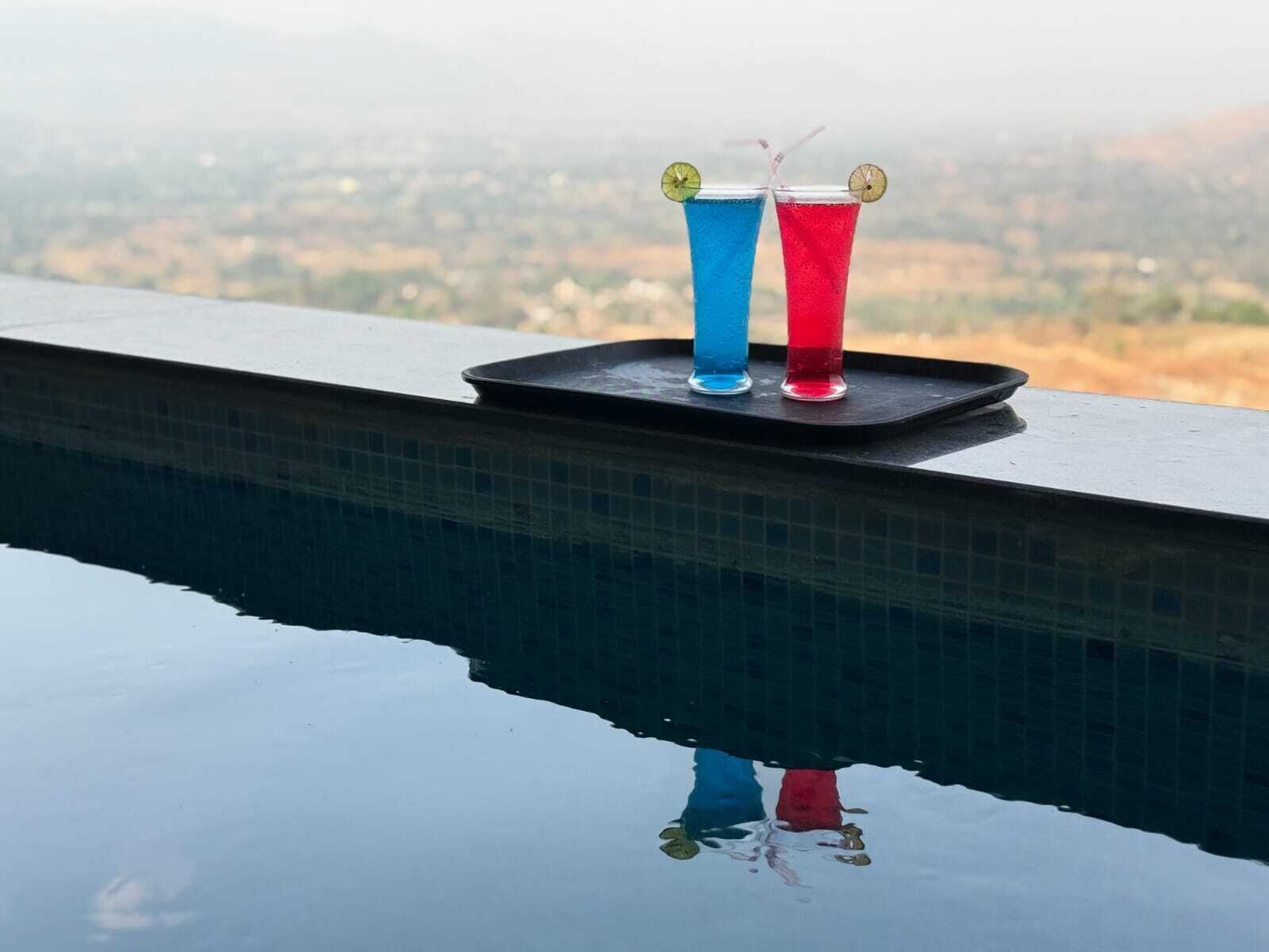 Deux verres d’une boisson bleue et rouge sont posés sur un plateau près d’une piscine, avec vue sur un paysage lointain.