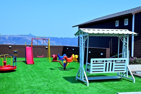 A playground with swings, slides, and climbing structures, a white bench, and a building with a sloped roof.