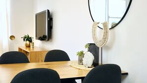 Een eetruimte met een houten tafel, zwarte stoelen, een aan de muur bevestigde televisie en een ronde spiegel met een gevlochten lijst.