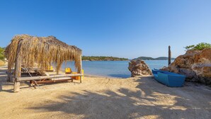 A beach with a thatched structure, a wooden bench, a blue boat, and a rocky outcrop. - Khaja Beach Hotel (Ayvalik)
