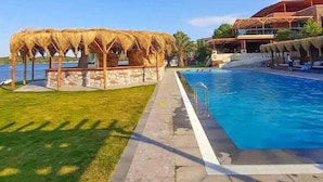 A swimming pool with a thatched structure and a building in the background. - Khaja Beach Hotel (Ayvalik)