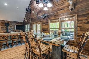 Un coin repas rustique avec des chaises en bois, une table en verre et une vue sur l’extérieur grâce à de grandes fenêtres.