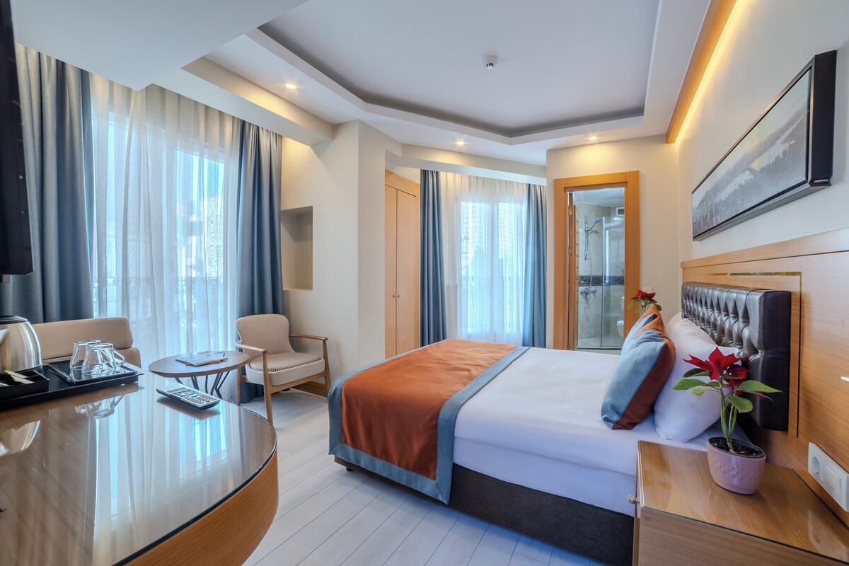 Una habitación de hotel con cama, mesita de noche, silla, escritorio y un gran ventanal con cortinas.