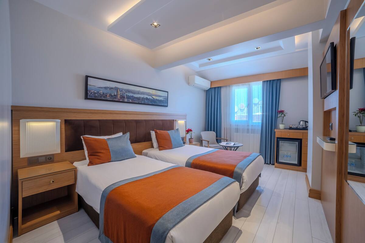 Habitación de hotel con dos camas, un escritorio, una silla, un televisor y una pintura de un paisaje urbano.