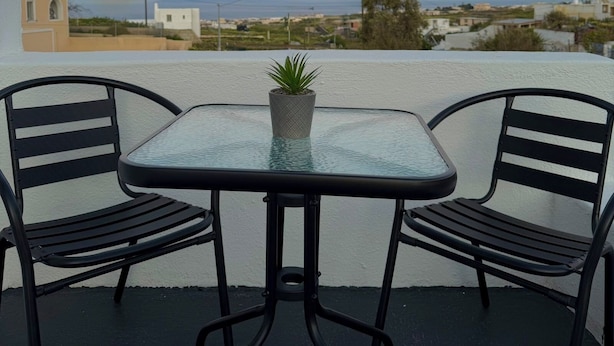 A balcony with a table and two chairs, overlooking a coastal landscape during sunset. - Raeti Fira Residence (Santorini)