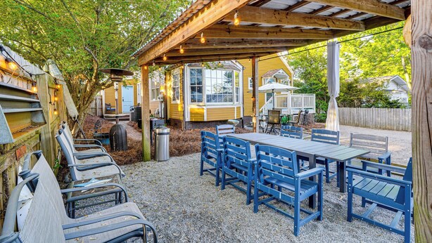 Ett takförsett utomhusområde med bord och stolar, en pergola med ljusslingor och en utsikt över ett hus i bakgrunden.