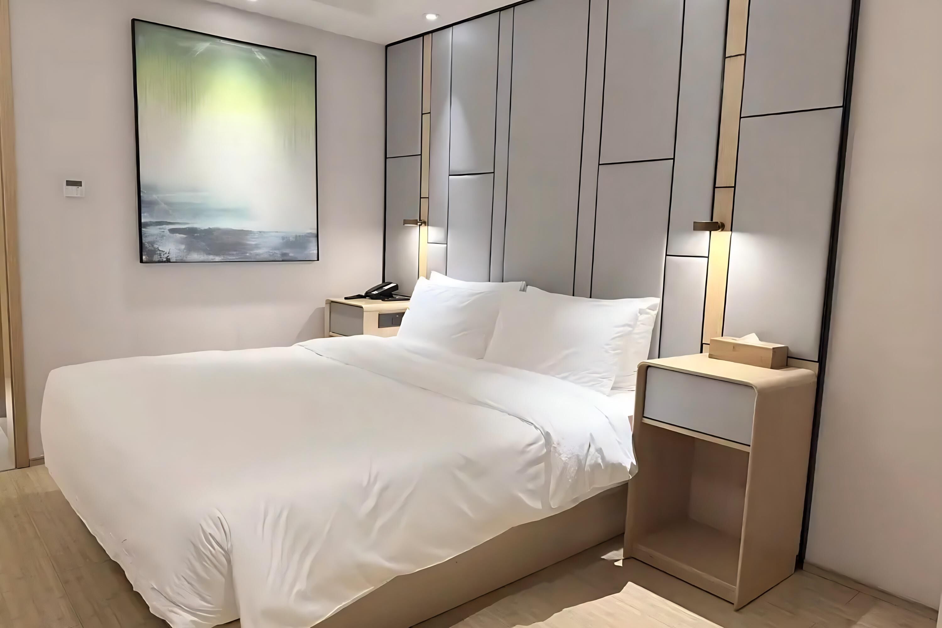 Una habitación de hotel moderna con una cama grande, mesitas de noche, una lámpara y una obra de arte colgada en la pared, además de un cabecero integrado.