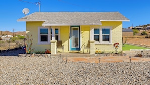 A single-story yellow house with a blue door, two windows, a satellite dish, and a small garden with flowers and a lawn. - Route 66 Roadtripper Retreat w/ Private Backyard (Kingman)
