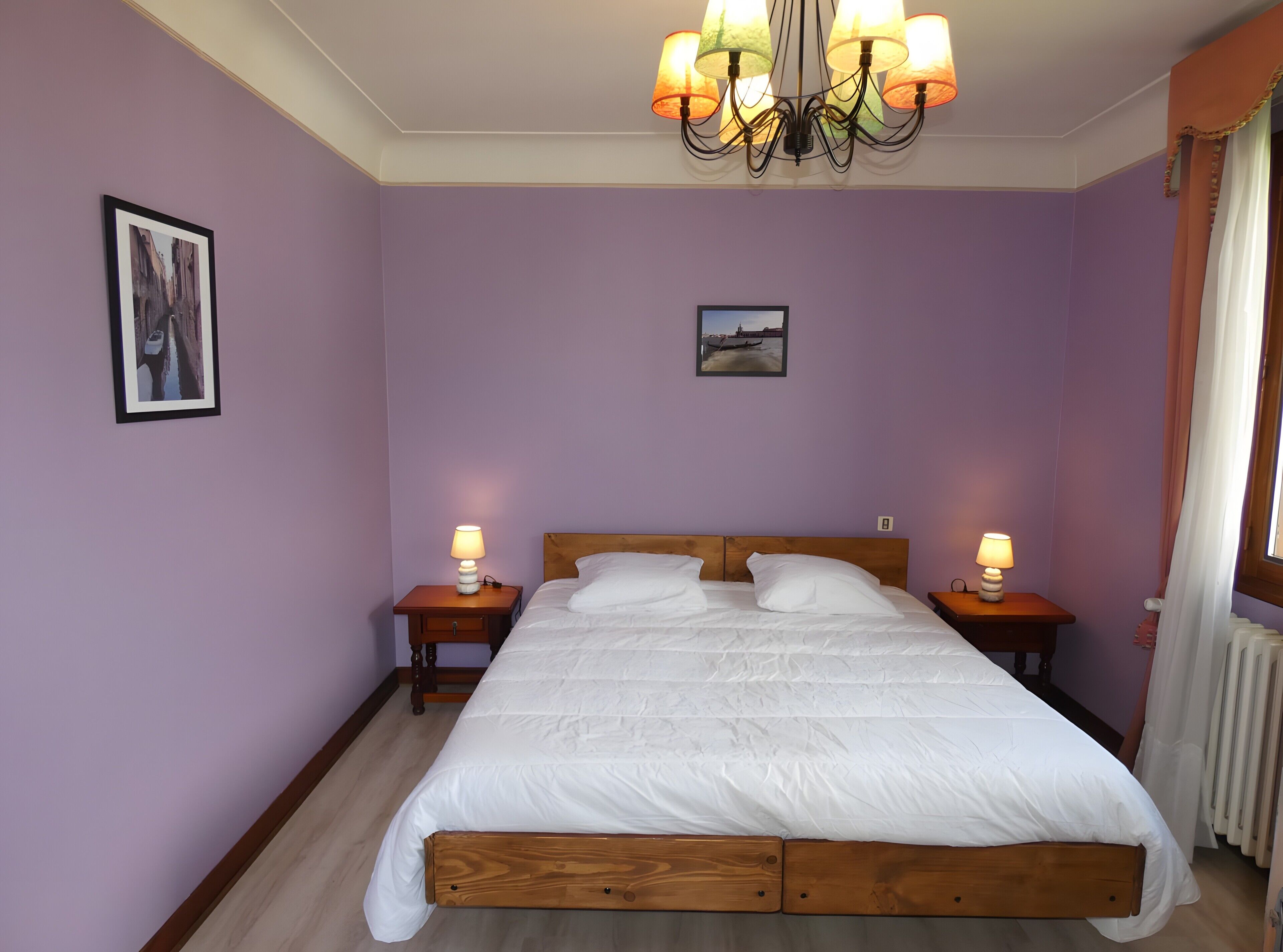 A bedroom with a wooden bed, two bedside tables with lamps, a chandelier, and two framed pictures on the walls.