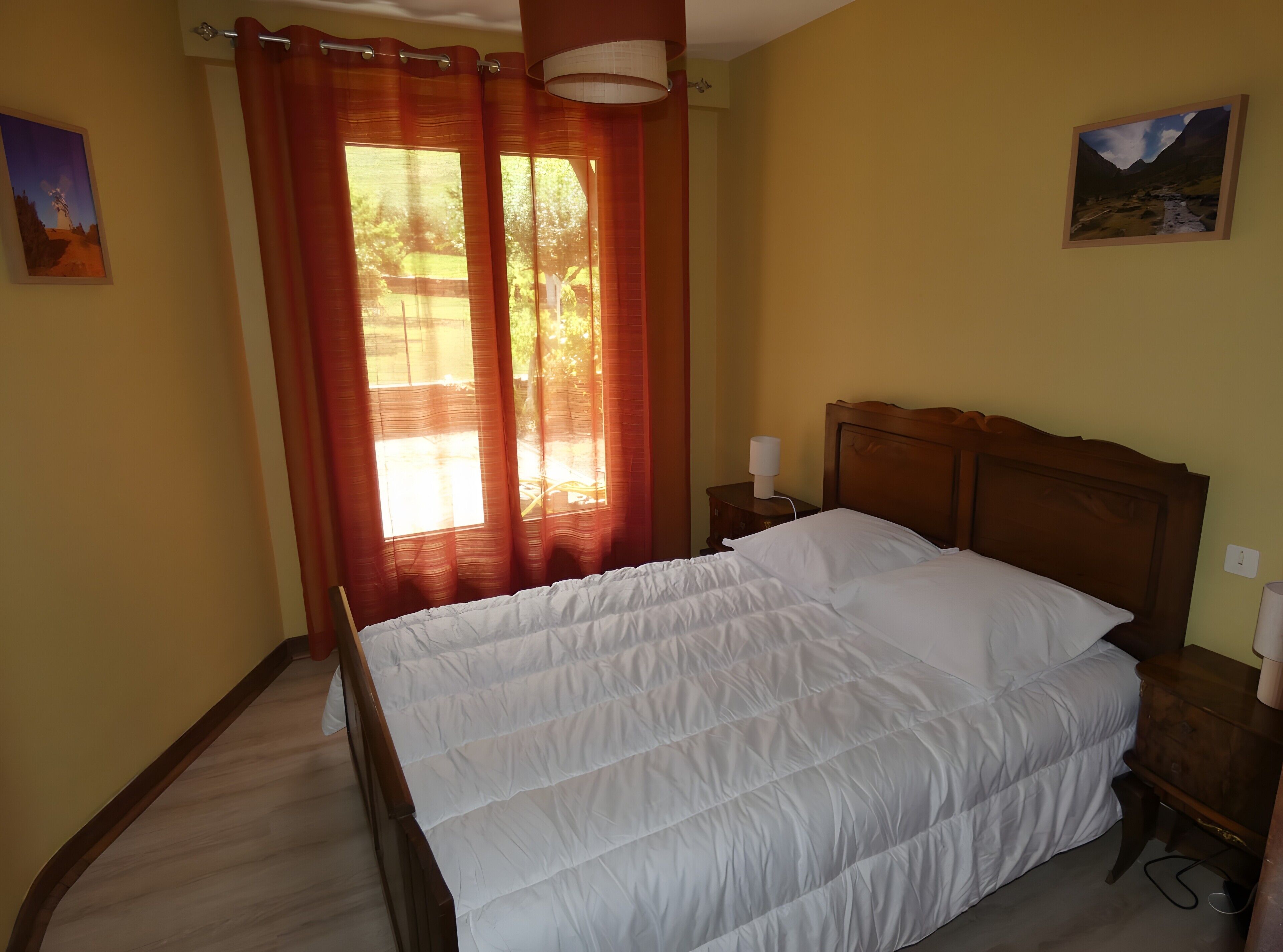 Un dormitorio con cama, cortinas, una mesita de noche, una lámpara y dos cuadros en la pared.