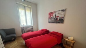 Ein Schlafzimmer mit zwei Betten, einem Sessel, einem kleinen Tisch, einem Fenster mit Jalousien und einem Bild an der Wand.