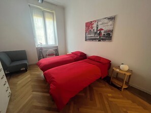 Ein Schlafzimmer mit zwei Betten, einem Sessel, einem kleinen Tisch, einem Fenster mit Jalousien und einem Bild an der Wand.