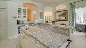 A kitchen with granite countertops, a double sink, and a dining area with a wicker sofa. - Gated Home w/ Pool, On-Site Golf in Port Charlotte (Port Charlotte)