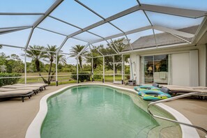 Ein überdachter Pool mit Liegestühlen und Luftmatratzen, umgeben von Palmen und einem Haus.
