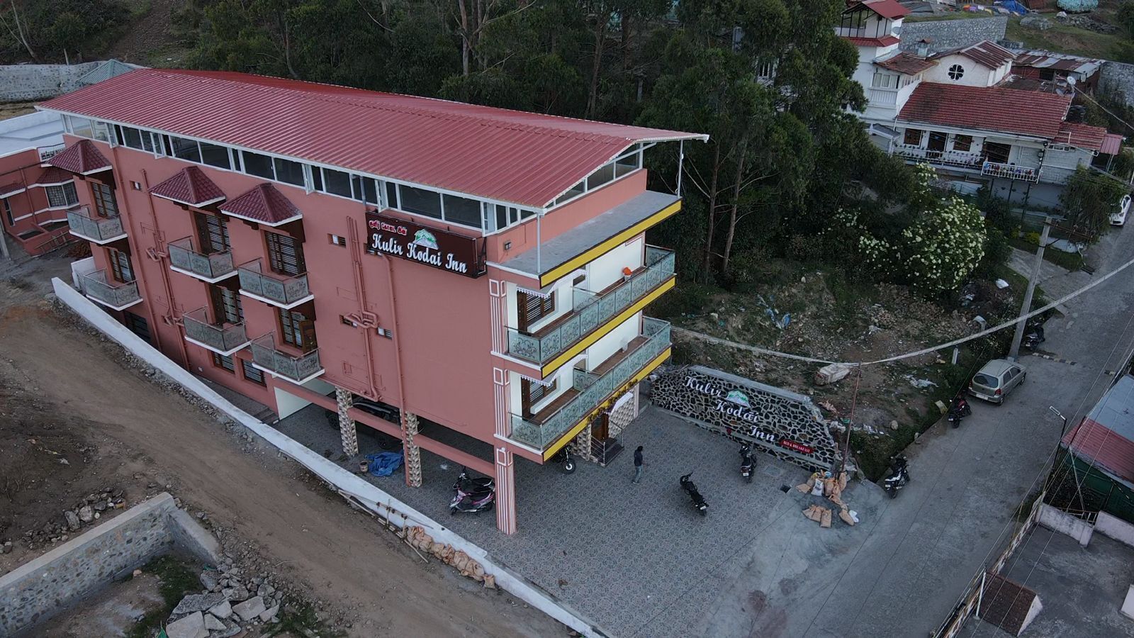 A multi-story building with balconies, a sign reading 'Kulir Kodai Inn,' and a paved ground area with parked vehicles.
