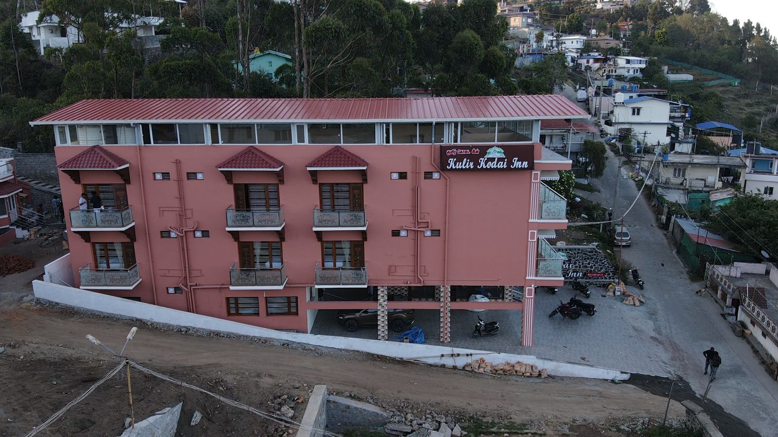A pink building with balconies, a sign reading 'Kulir Kodai Inn,' and a street view with parked vehicles and a person walking.