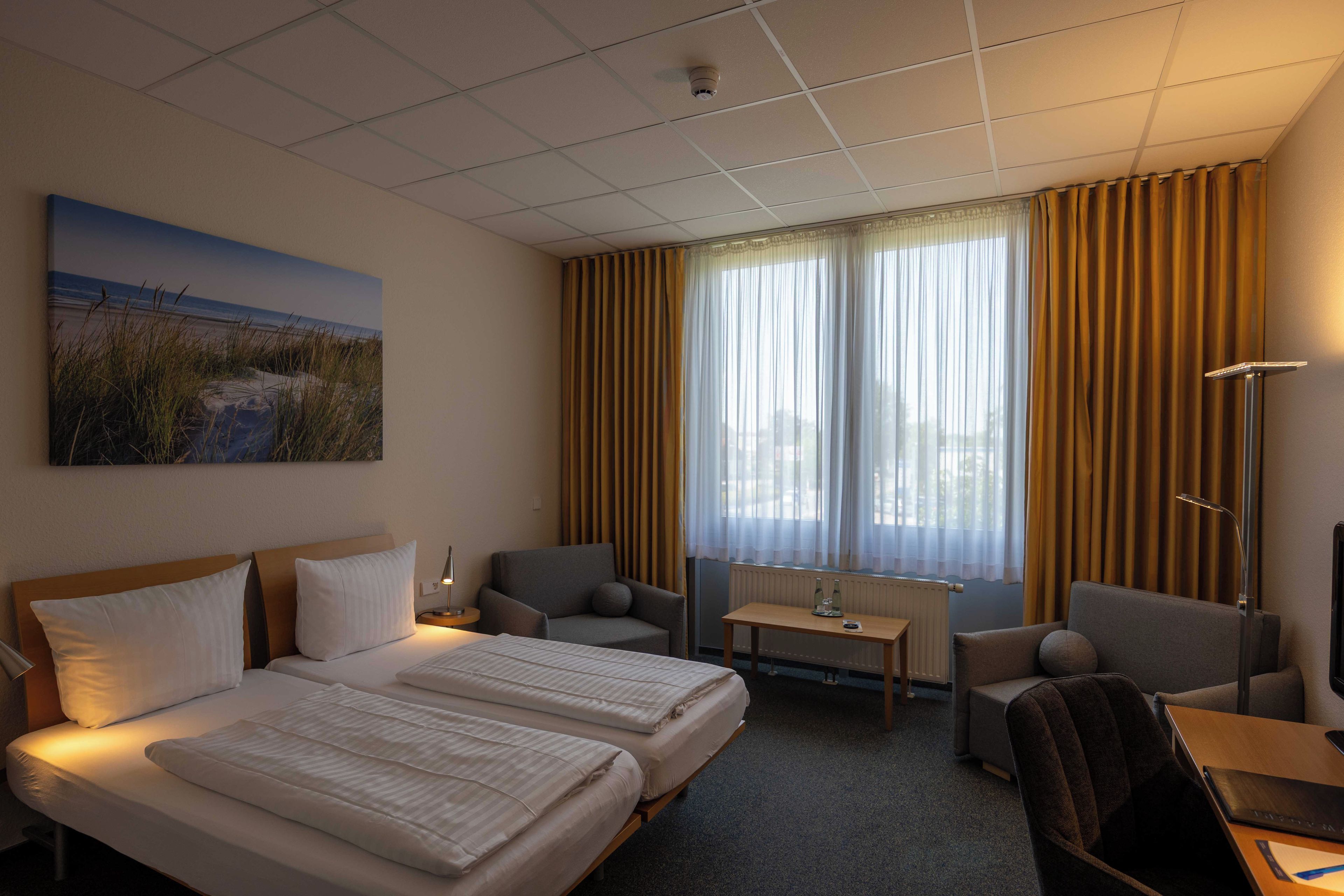 A hotel room with two beds, a desk, a chair, a lamp, and a painting on the wall.