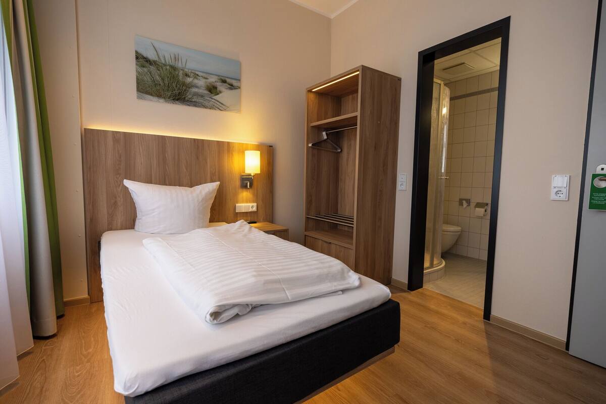 A neatly made bed in a hotel room with a wooden headboard, a bedside lamp, a wardrobe, and a view of the bathroom.