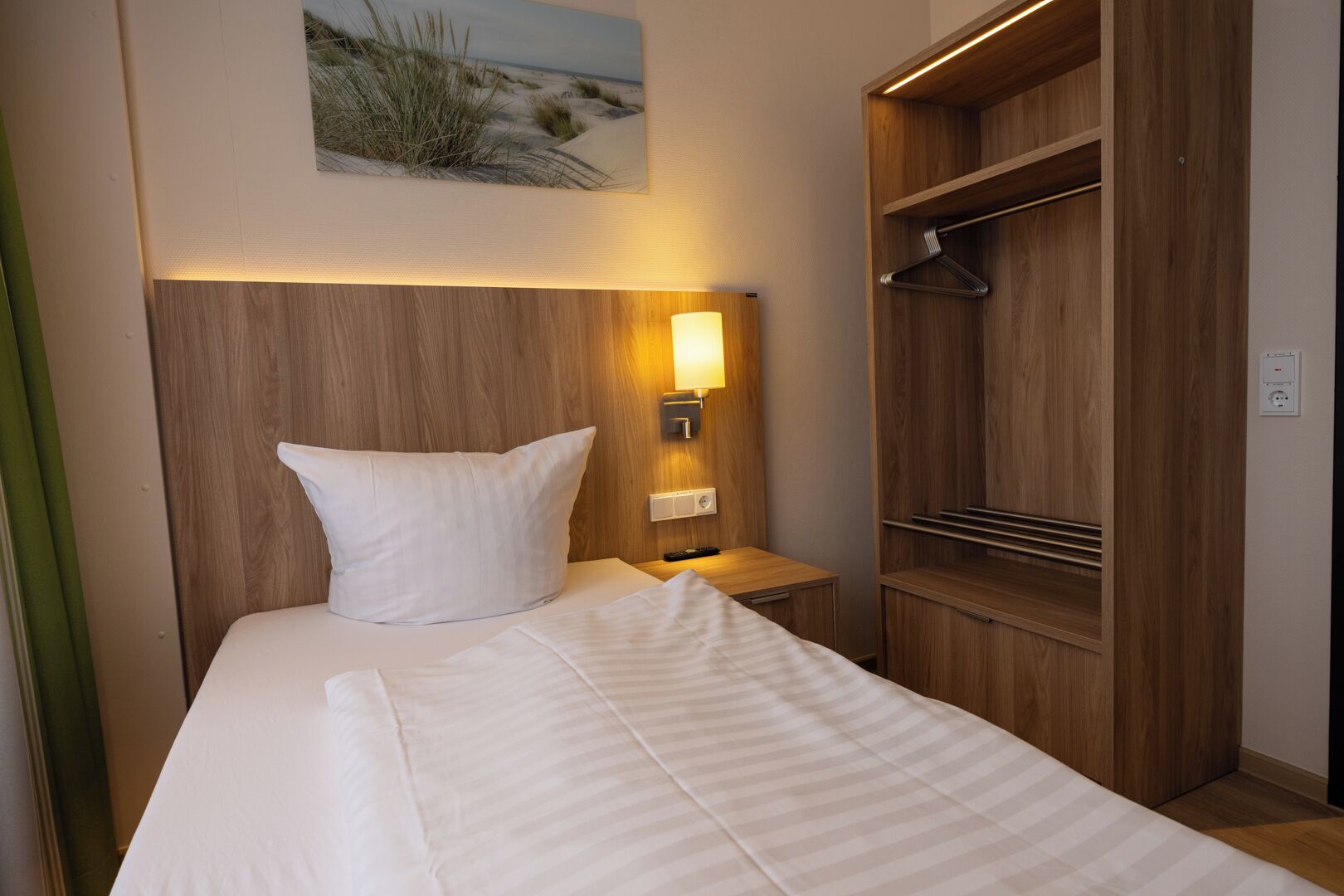 Ein Hotelzimmer mit Bett, Nachttisch, Wandlampe, Holzschrank und einem Strandbild.