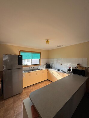 A kitchen with a refrigerator, stove, microwave, and a window with blinds. - Cosy Beach House, 10 mins walk to beach, pet friendly (Venus Bay)