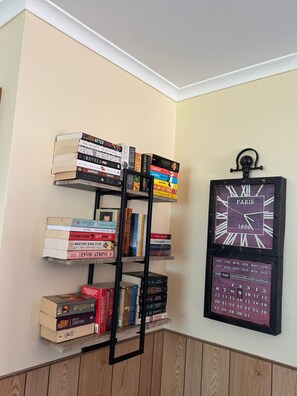 A corner bookshelf with a variety of books and a wall-mounted clock. - Cosy Beach House, 10 mins walk to beach, pet friendly (Venus Bay)