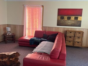 Un sofá de cuero rojo, una mesita de madera, un mueble de almacenamiento de mimbre y una ventana con cortinas traslúcidas.