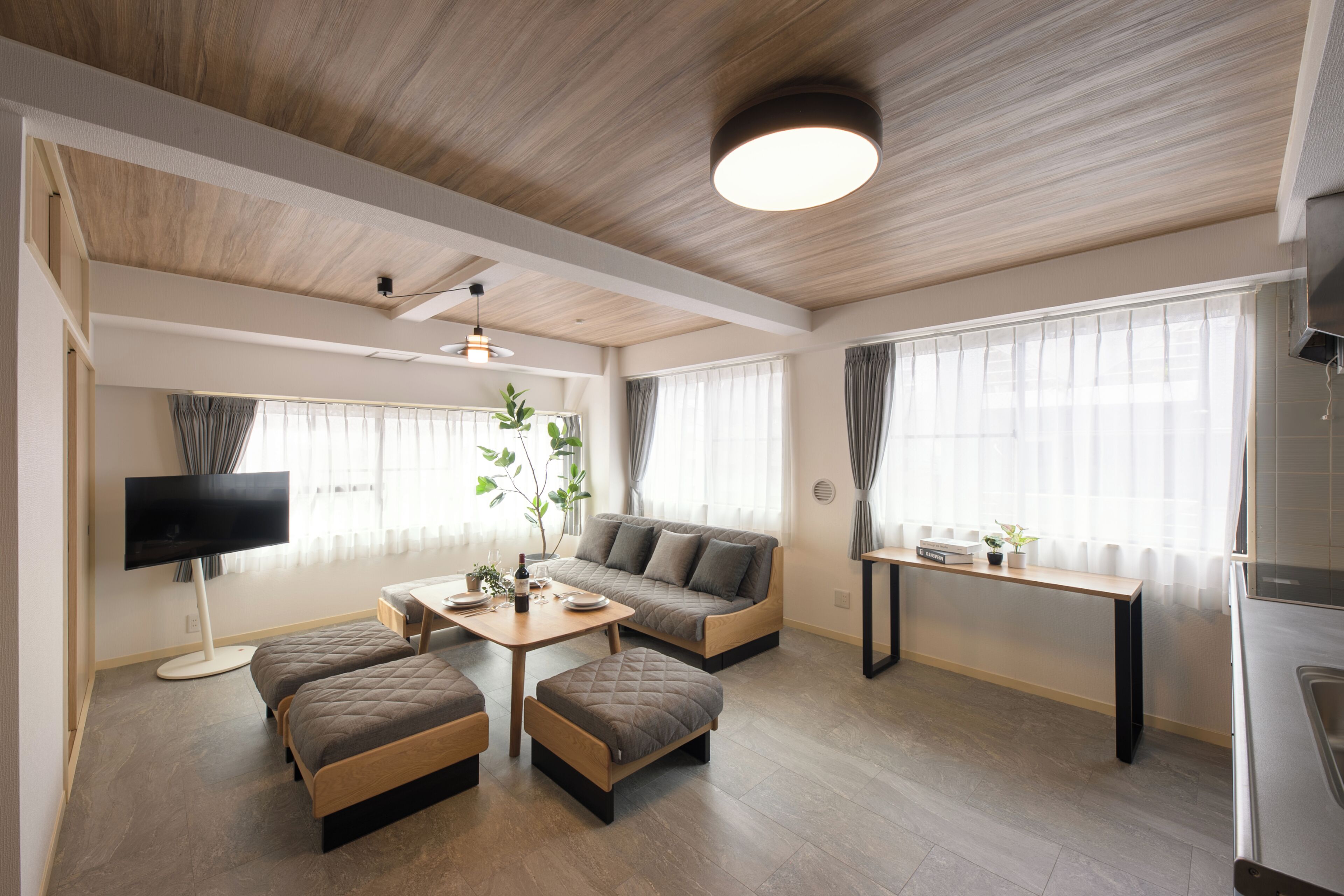 A modern living room with a sofa, ottomans, a TV, a dining table, and a kitchen area.