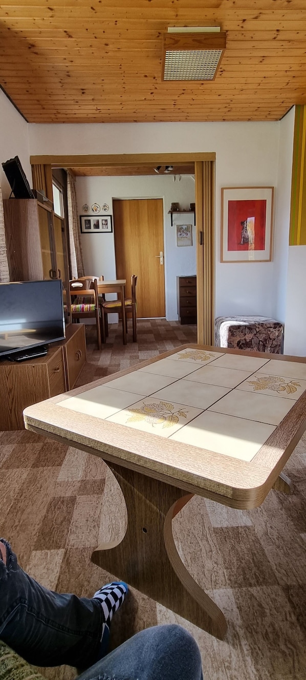 A room with a wooden ceiling, a large table with a floral tile design, a TV on a wooden cabinet, and a door leading to another room.
