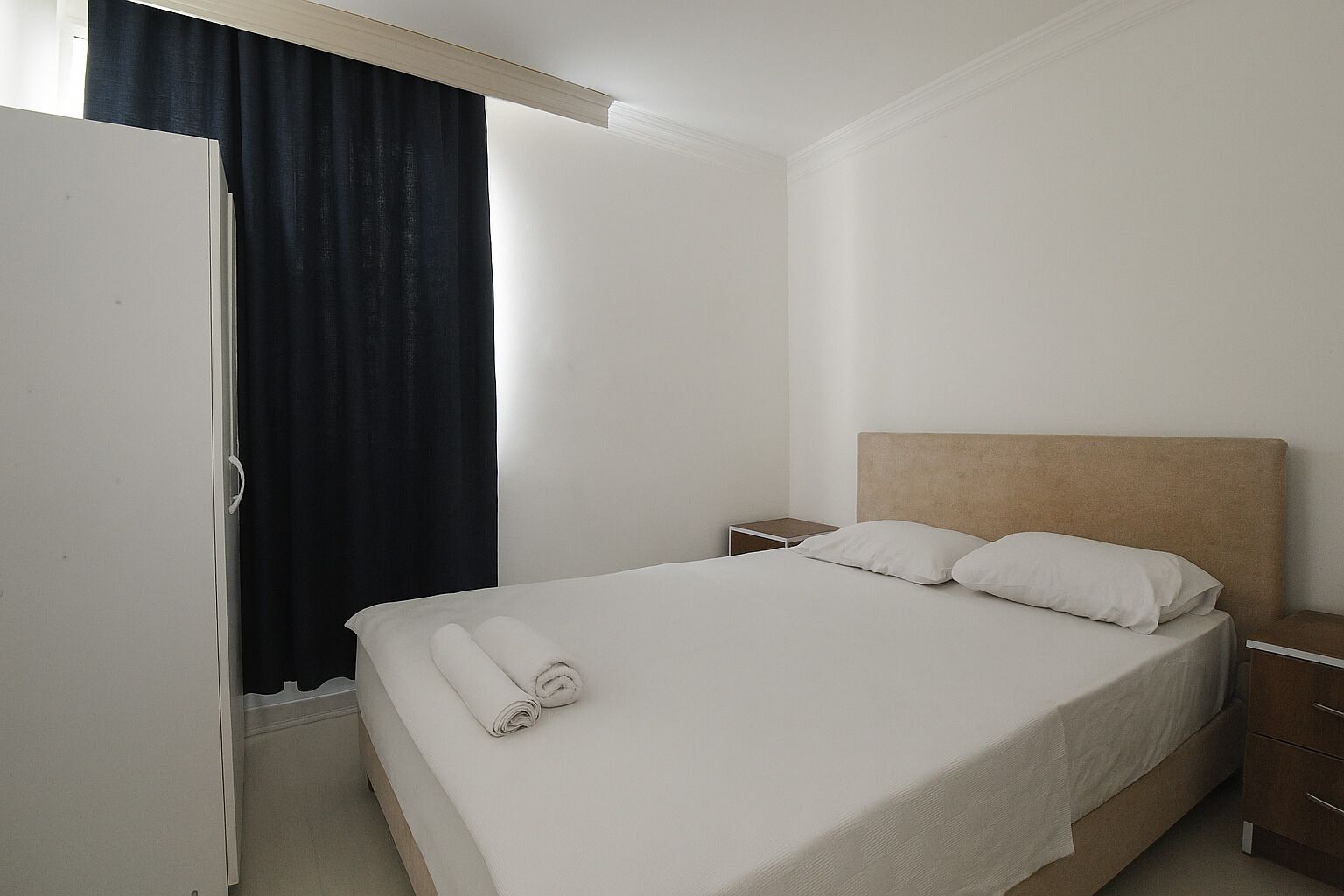 Una cama individual con ropa blanca, cabecera de madera, mesita de noche y cortina oscura.