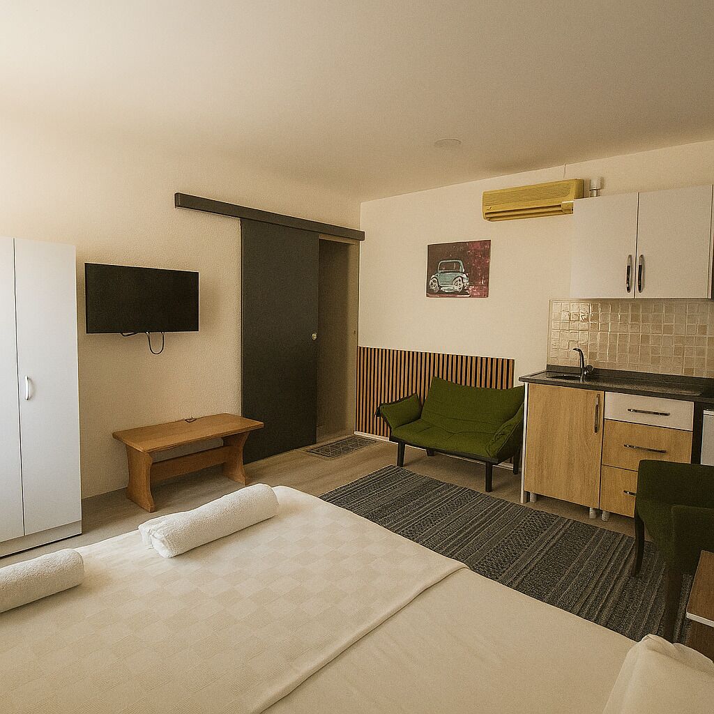 A bedroom with a bed, a bedside table, a bench, a TV, a kitchenette, and a door.