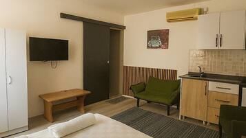 A bedroom with a bed, a bedside table, a bench, a TV, a kitchenette, and a door.