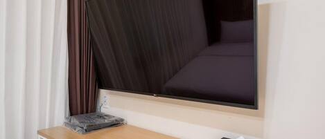 A hotel room with a flat-screen TV mounted on the wall, a wooden desk with a tissue box, and a bed with dark bedding.