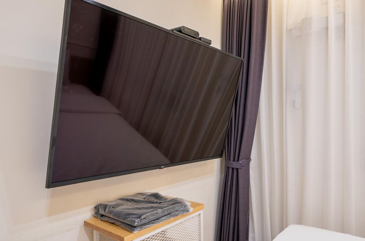 A room with a large flat-screen TV mounted on the wall, a bed with dark bedding, a small table with a folded towel, and a white curtain.