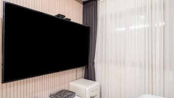 A hotel room with a large flat-screen TV mounted on the wall, a small white bedside table with a tissue box and a magazine, a white bedside unit, and sheer white curtains.